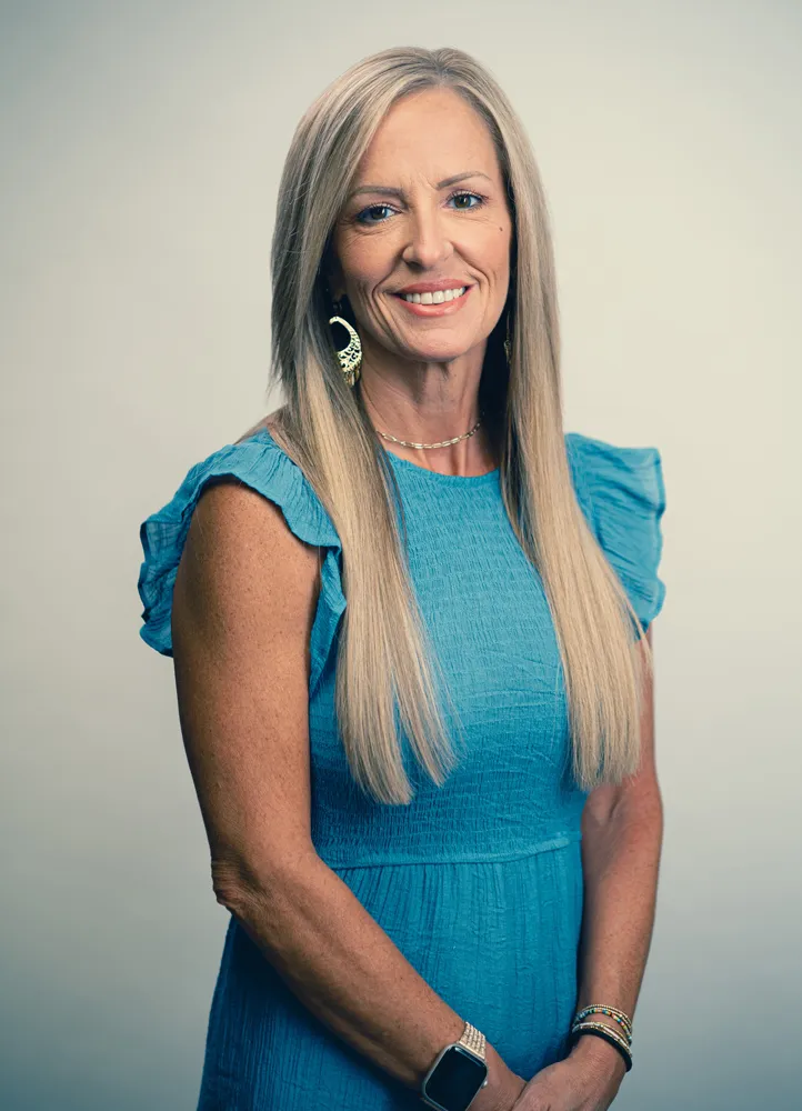 Professional headshot of a smiling woman in blue dress with long blonde hair, earrings and smartwatch on neutral background