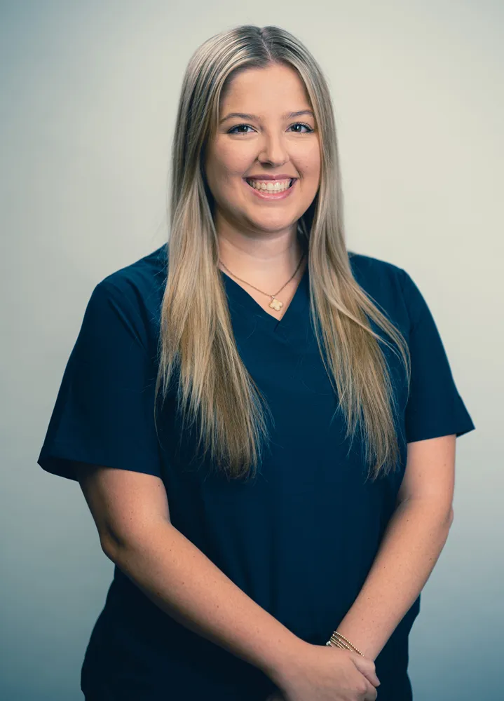 Smiling woman in navy medical scrubs, long blonde hair, professional headshot on neutral background.