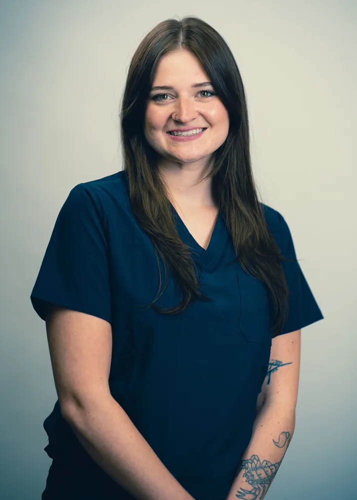 Smiling woman in navy medical scrubs, long brown hair, visible forearm tattoo, studio portrait on neutral background