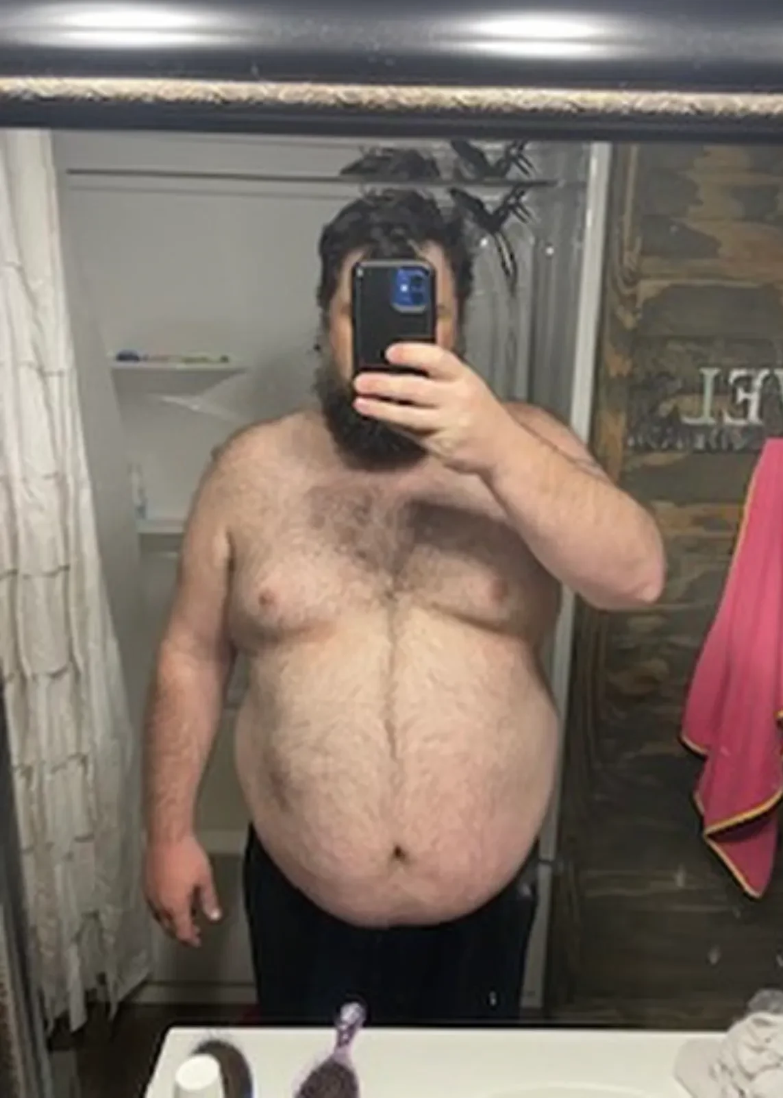 A bearded man taking a bare-chested bathroom mirror selfie with smartphone, visible chest and belly