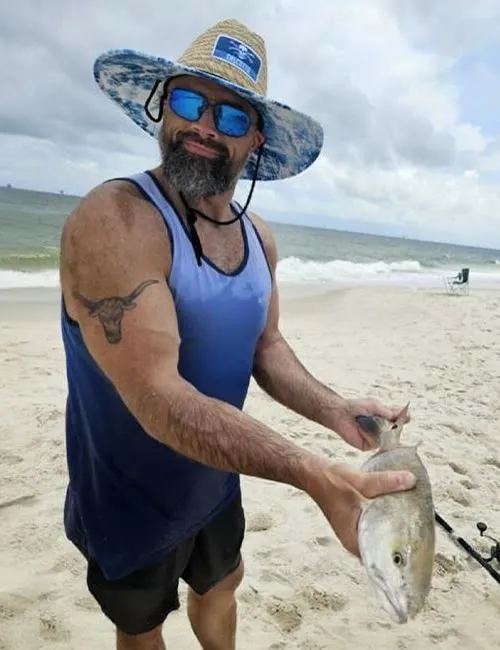 Man in sunhat and sunglasses holding a freshly caught fish on a sandy beach, showing an arm tattoo and fishing gear