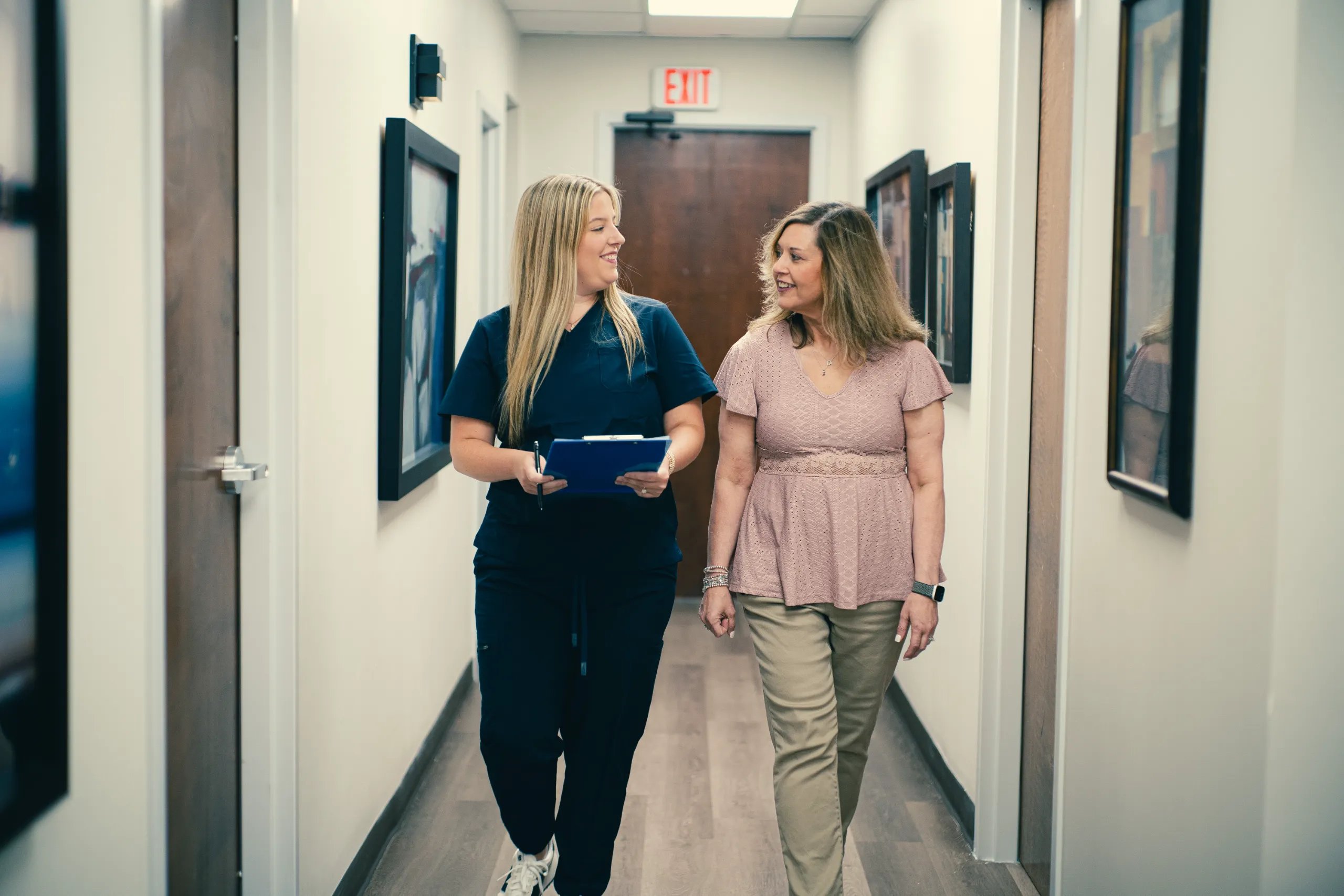 Two women walking in a clinic hallway, woman in scrubs with clipboard speaking to another smiling woman
