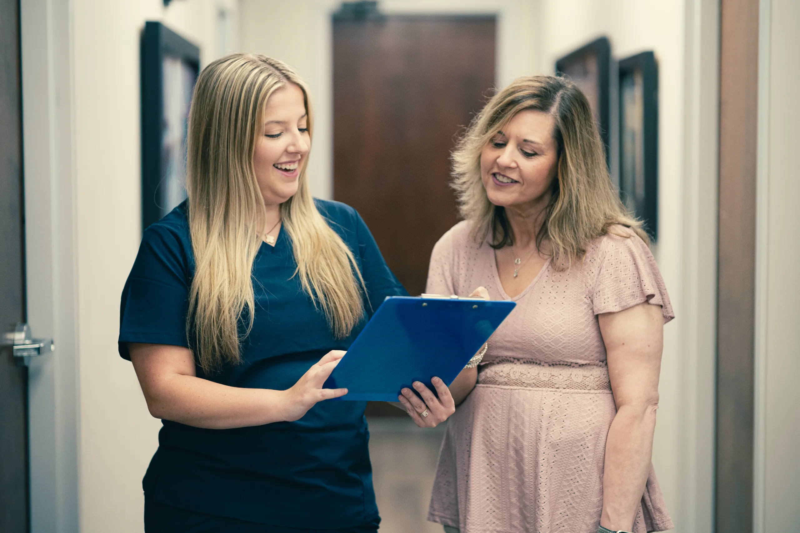 A woman in navy scrubs reviews a blue clipboard with a smiling patient in a clinic hallway