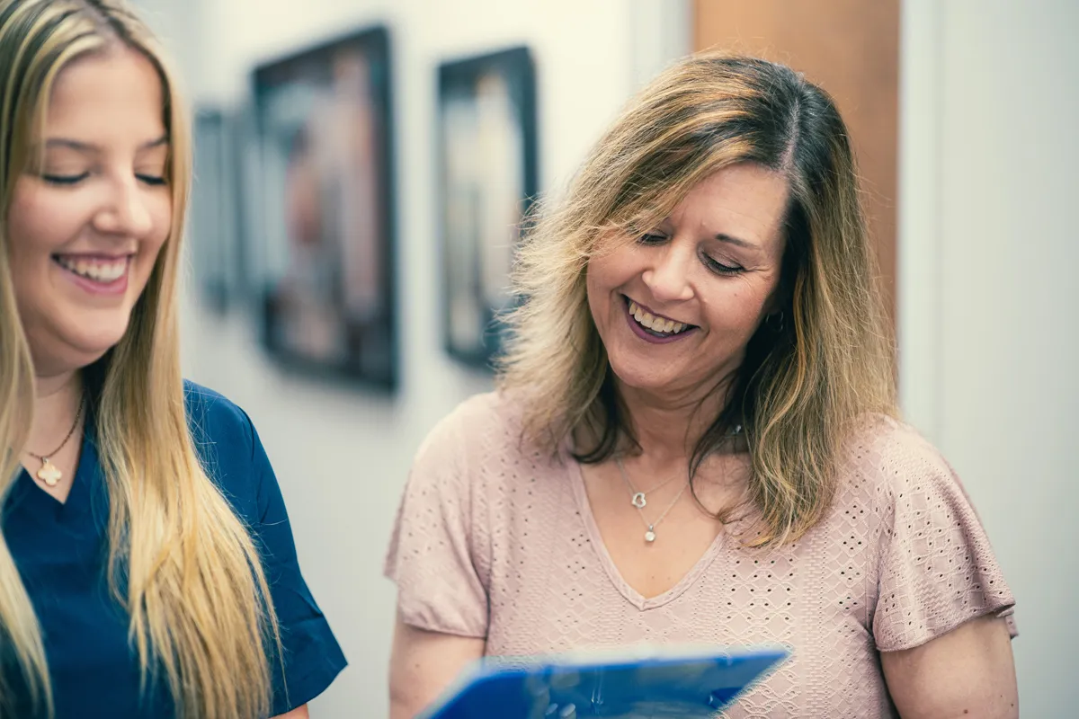 Two women smiling while reviewing a blue clipboard in a bright office hallway