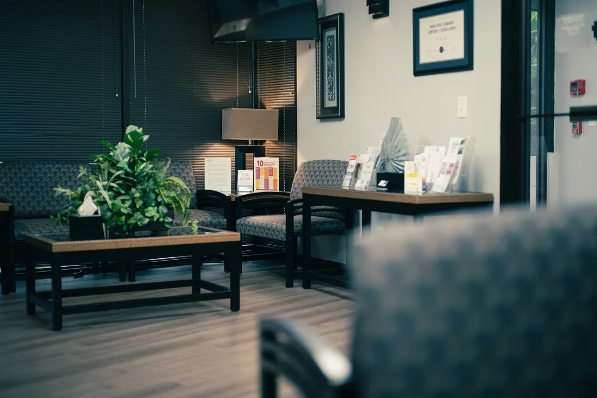 Medical office waiting room with patterned chairs, wooden tables, green potted plant, brochures and a lamp.