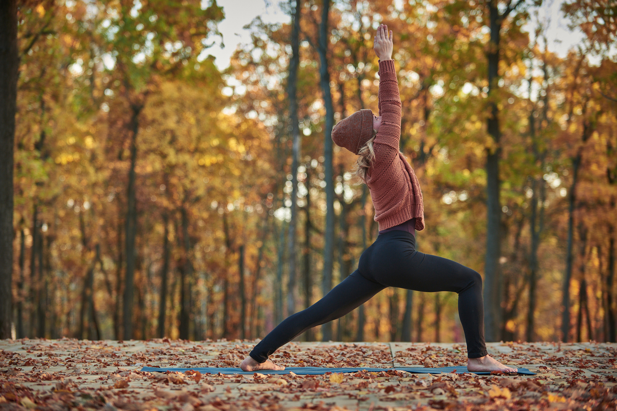 A woman in layers doing yoga outside among colorful autumn trees.